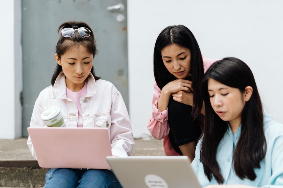 Three young Asian women working together on laptops outdoors, highlighting education and technology.
